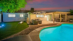 A backyard with a swimming pool, a covered patio, and a lawn.