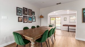A dining area with a wooden table and green chairs, a wall with framed artwork, and a doorway leading to an outdoor patio.