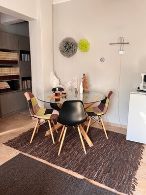 Un coin repas avec une table en verre, des chaises noires et multicolores, un tapis marron, une bibliothèque et un meuble blanc.
