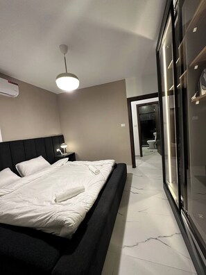 A modern hotel room with a large bed, bedside table, lamp, and an air conditioning unit. - Stylish Luxe Apartment Fr 0003 (Amman)