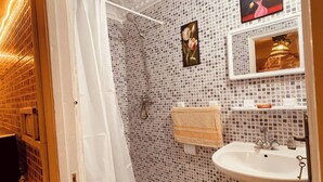 Et badeværelse med hvid toilet, vask, bruseniche med bruseforhæng og en flisevæg med et blomsterbillede.