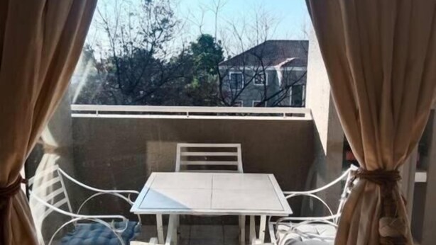 Un balcón con mesa y sillas, cortinas y vistas a árboles y edificios.