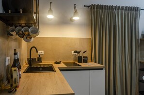 Una cocina con encimera de madera, pileta negra, tazas colgantes, dos lámparas colgantes y una cortina.
