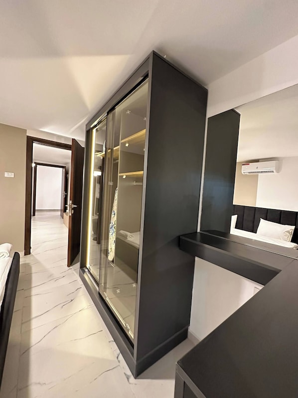 A modern hotel room with a large sliding glass door closet, a bed with a black headboard, and a bedside table.