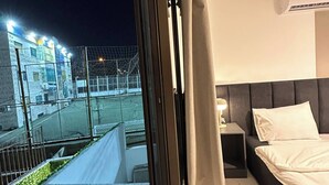 A bedroom with a bed, a nightstand, an air conditioning unit, and a view of a sports field through a window. - Stylish Luxe Apartment Fr 0006 (Amman)