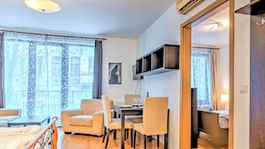 Ett modernt vardagsrum med trägolv, ett matbord med stolar, en soffa, en tv som är monterad på väggen och ett stort fönster med gardiner.