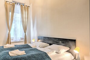 Ein ordentlich bezogenes Bett mit weißen Leinentüchern, einer blauen Decke und einem Kopfteil. Es gibt ein Fenster mit Vorhängen, eine Nachttischlampe und eine Wand-Klimaanlage.