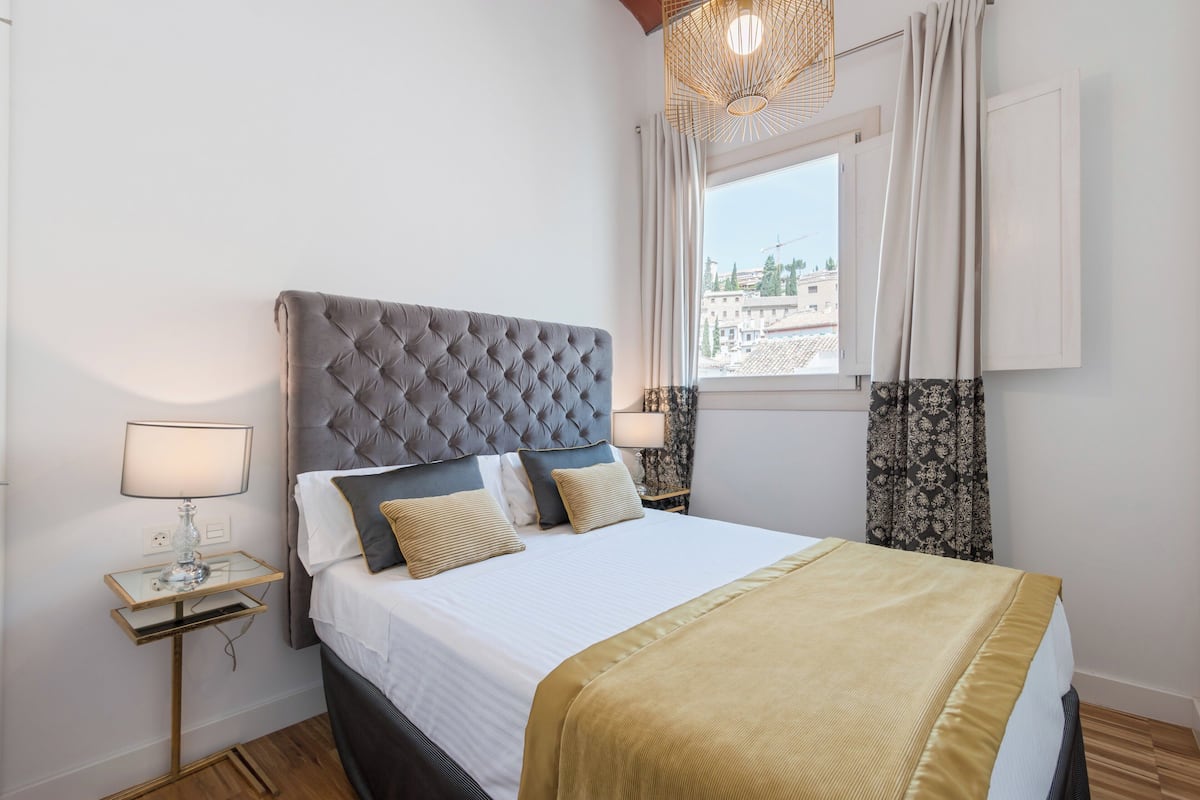 Un dormitorio ordenado con un cabecero capitonado, una cama grande con ropa de cama blanca y un plaid dorado, una mesita de noche con una lámpara y una ventana con cortinas.