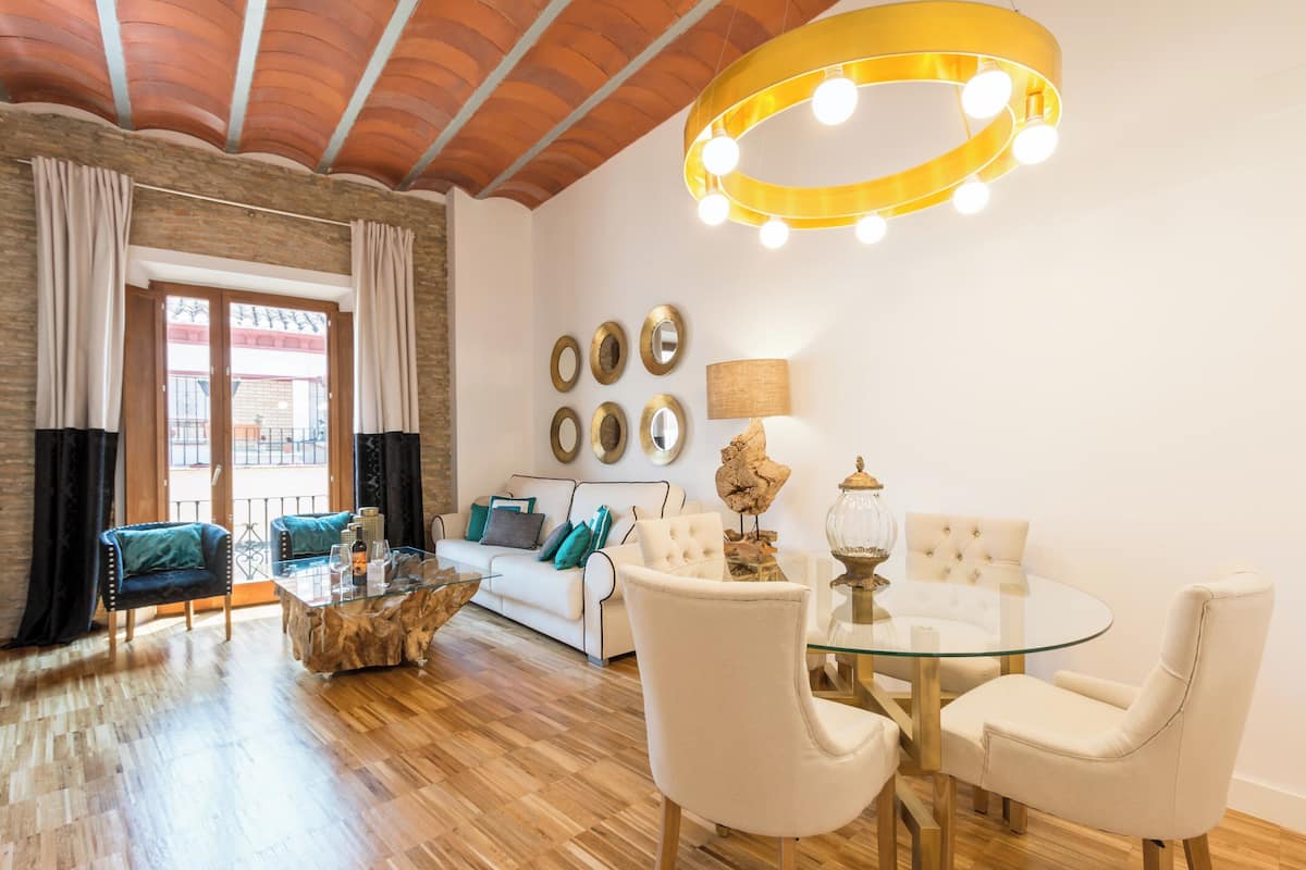 Amplia sala de estar con piso de madera, una mesa de centro de vidrio, un sofá blanco y una mesa redonda de vidrio con cuatro sillas. Sobre la mesa hay una lámpara circular grande y un conjunto de espejos enmarcados en la pared.