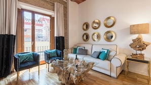 Un salón con una mesa de centro de vidrio, un sofá blanco con cojines azules, un sillón negro y una mesita de madera con una lámpara.