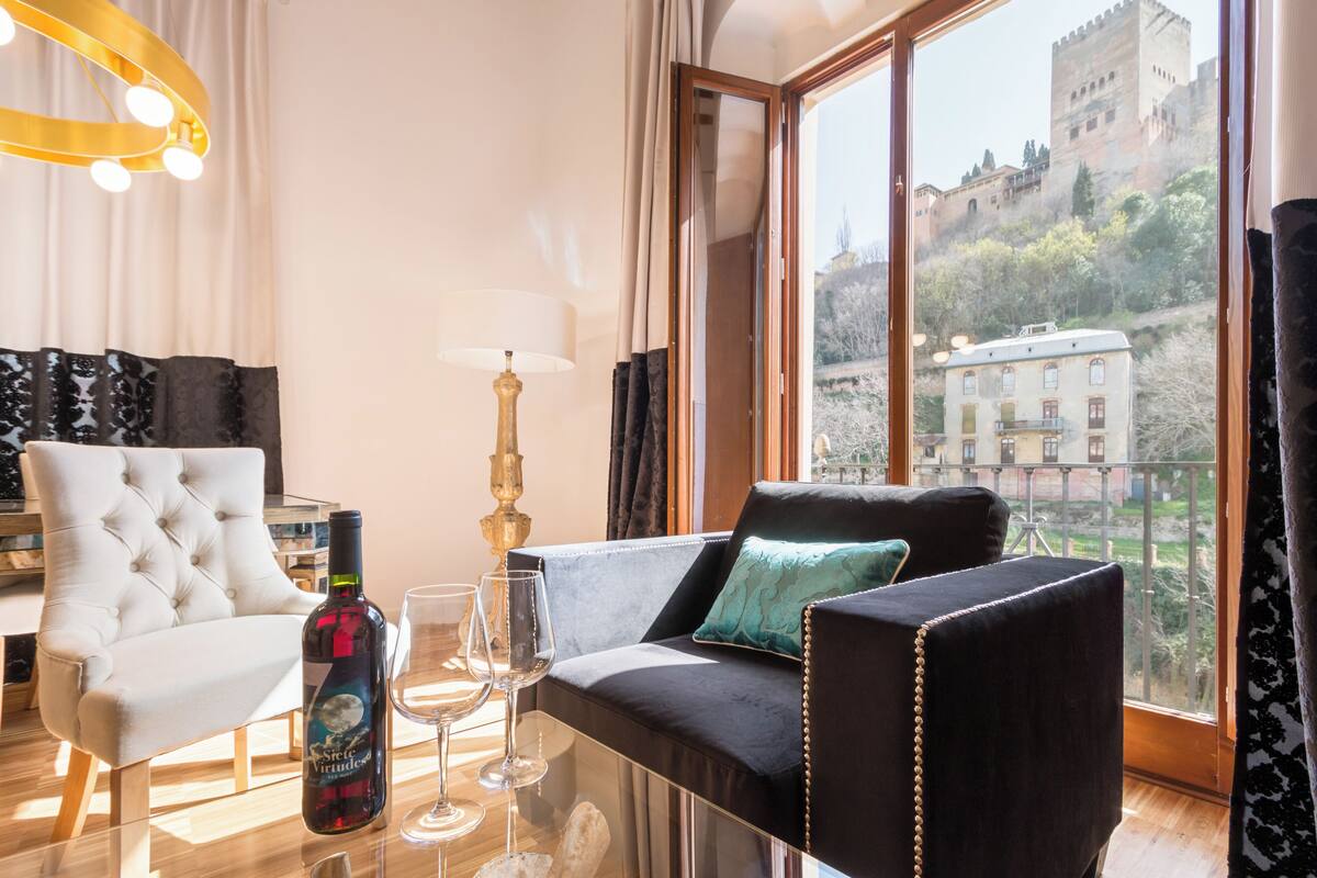 Una sala de estar moderna con un sillón blanco capitonado, un sofá negro con un cojín verde azulado, una mesa de vidrio con una botella y copas de vino, y vistas a un castillo a través de amplios ventanales.