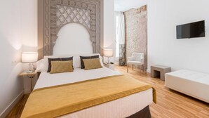Una habitación de hotel con una cama grande, dos mesitas de noche con lámparas, un televisor montado en la pared, un sofá blanco y una pequeña mesa.