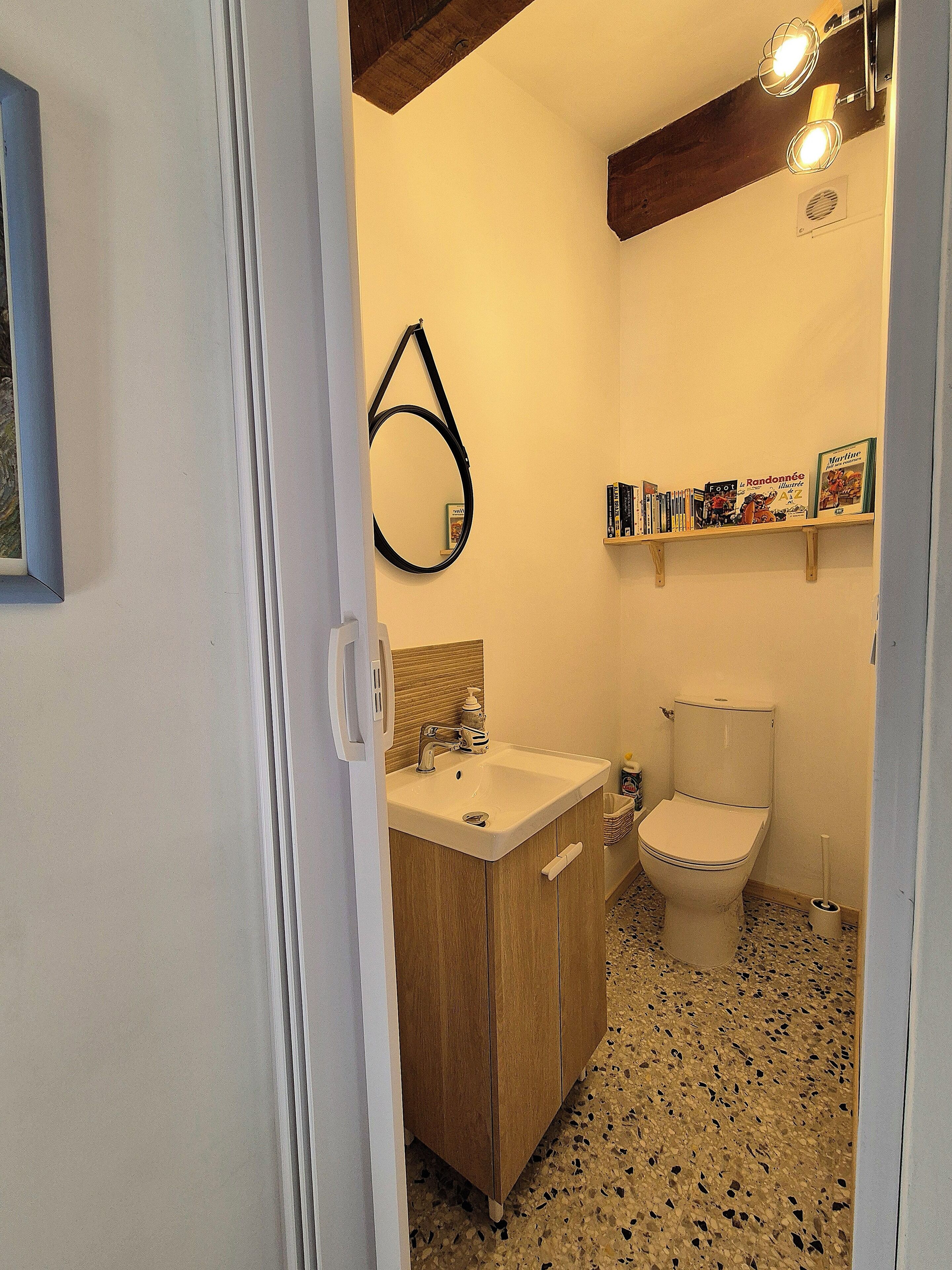 Une salle de bain avec un meuble en bois, un lavabo blanc et des toilettes. On y trouve une étagère avec des livres et un miroir accroché au mur.