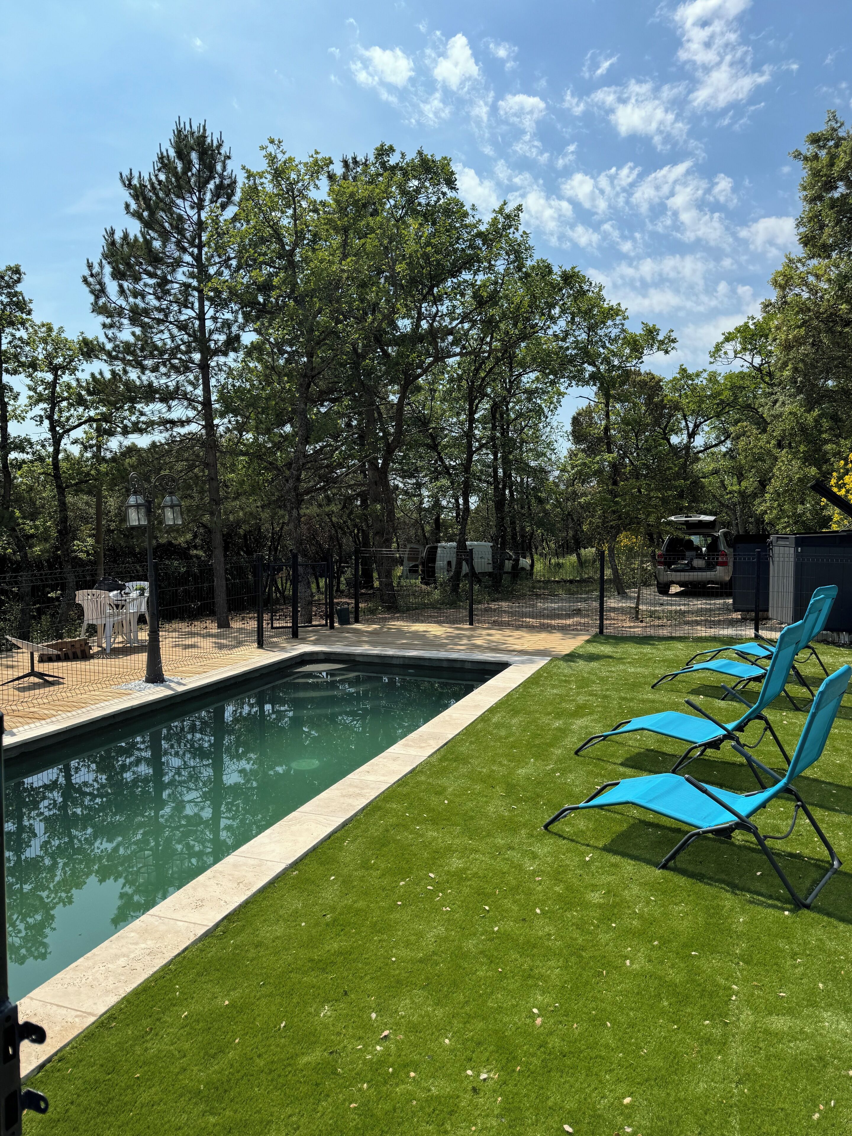 Une piscine entourée de pelouse verdoyante et d’arbres, avec des chaises longues et une zone délimitée par une clôture.