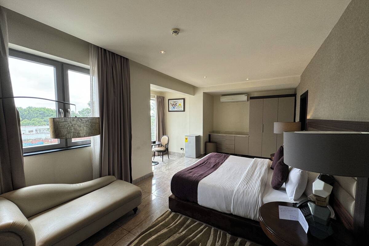 A modern hotel room with a bed, a sofa, a desk with a lamp, a chair, and a view of the outdoors.