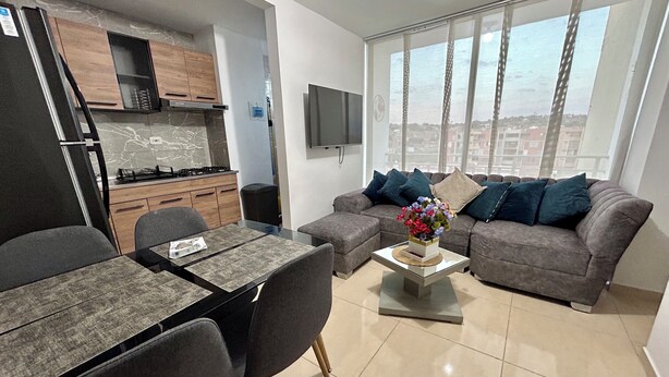 A modern living room with a grey sofa, a dining table with chairs, a kitchen area with wooden cabinets, and a large window with a city view. - Lindo apartamento 3 habitaciones (San José de Cúcuta)