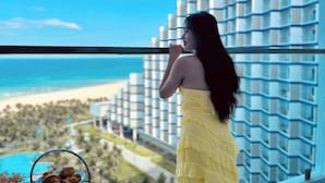 Una persona con un vestido amarillo de pie junto a un balcón con vista a una playa y edificios de hotel.