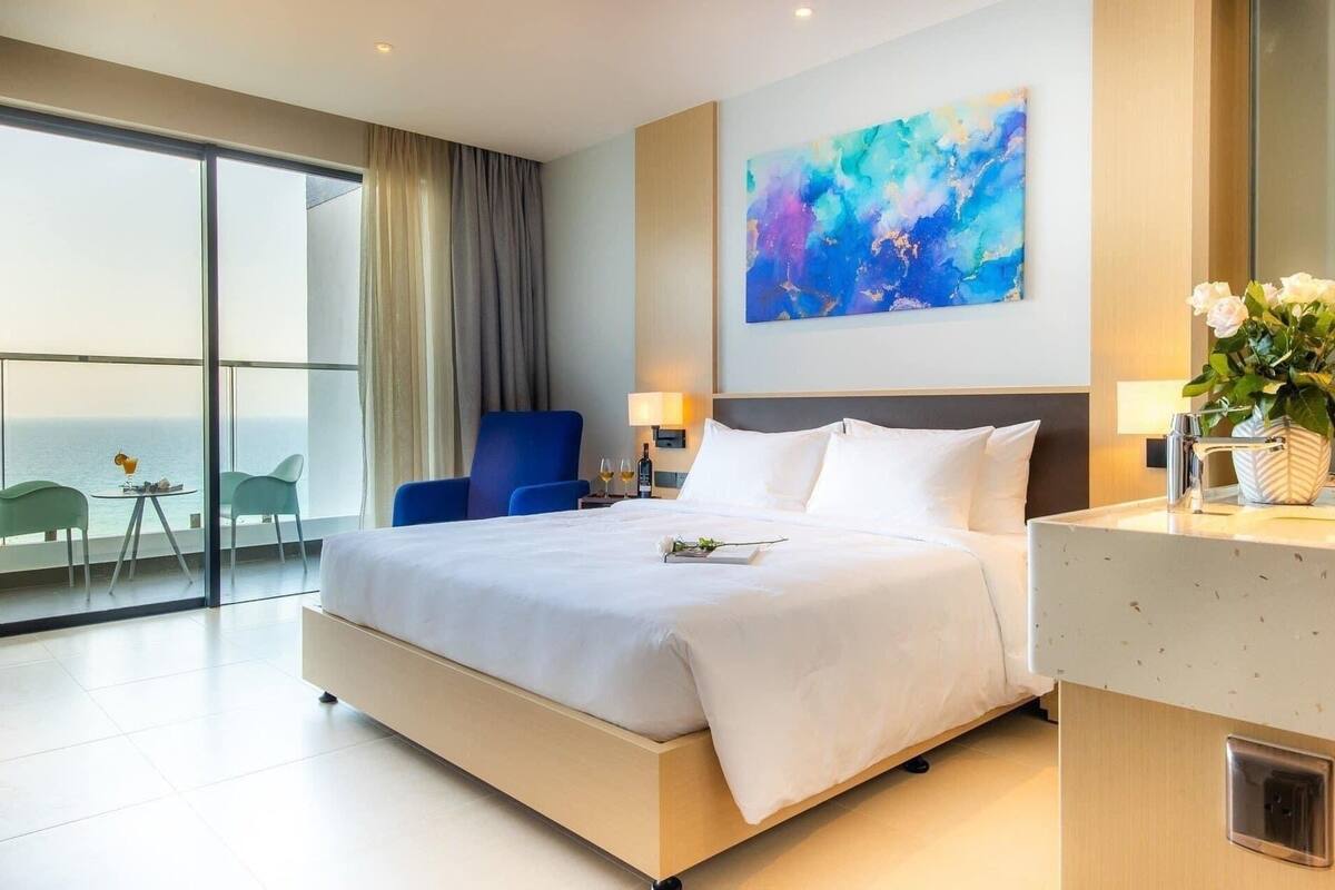 A modern hotel room with a large bed, a balcony with a view, a blue chair, and a painting on the wall.