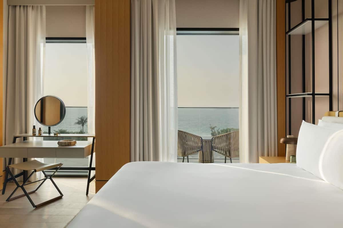 A hotel room with a large bed, a desk with a chair, a small table, a mirror, and a view of the ocean.