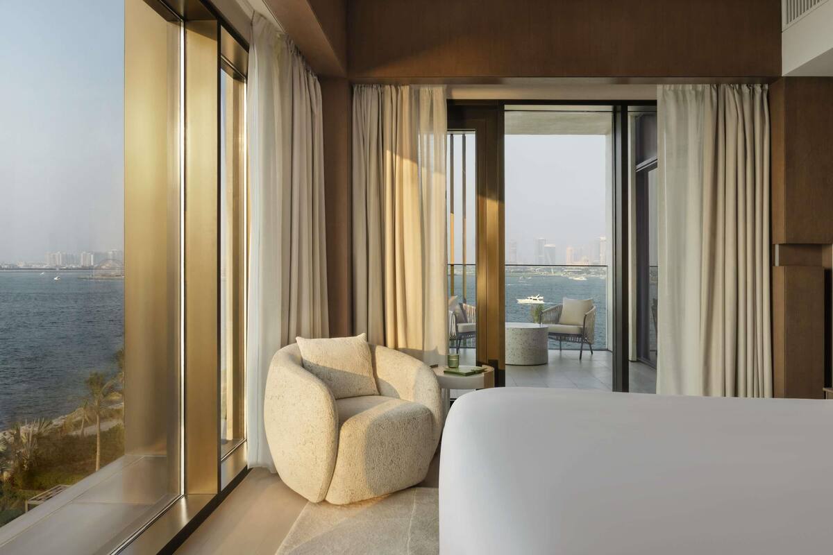 A modern hotel room with a large bed, a comfortable armchair, and a balcony with a view of the water and city skyline.