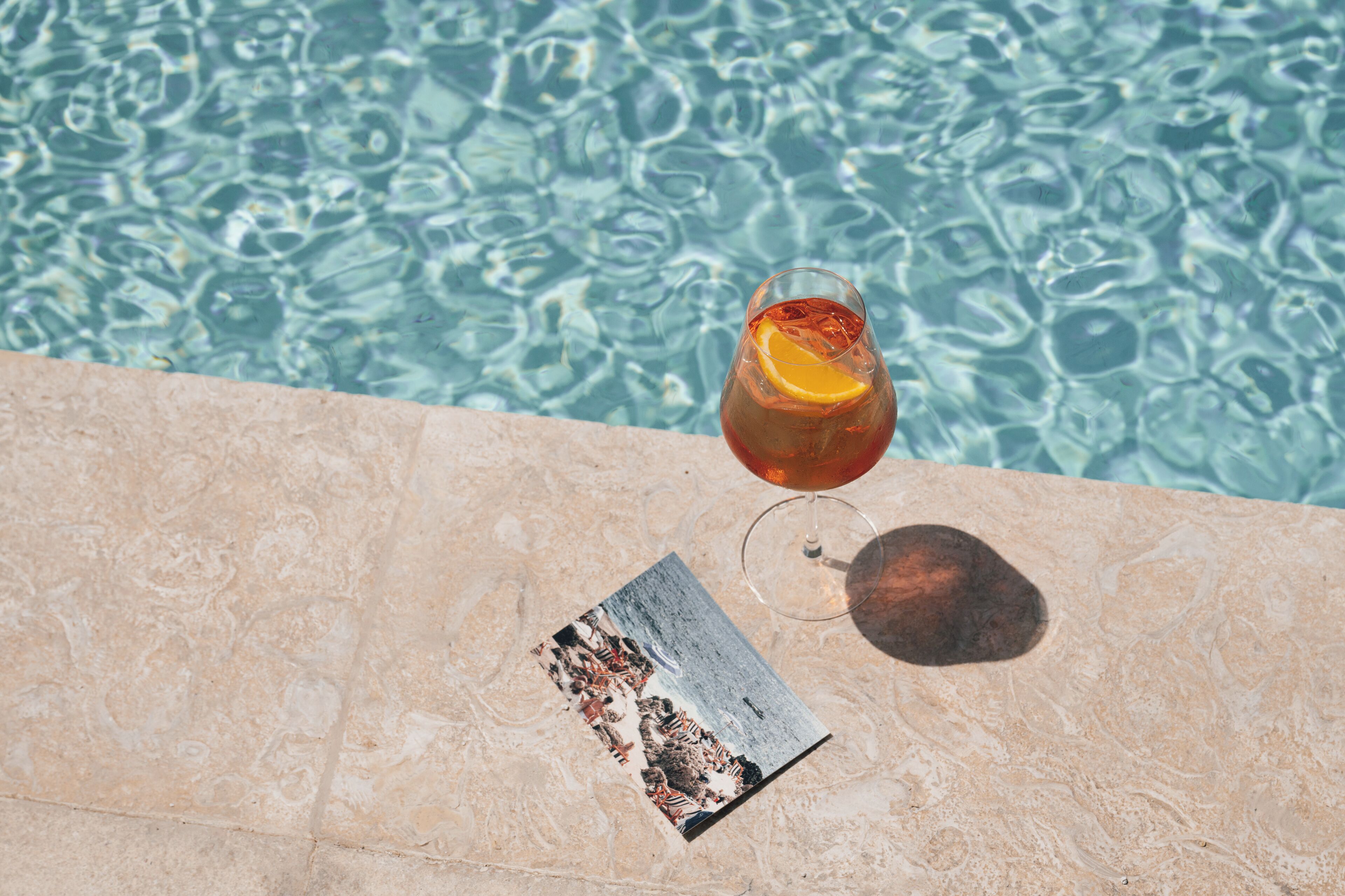Ein Getränk mit Orangenscheibe an einem Pool, daneben eine Postkarte.