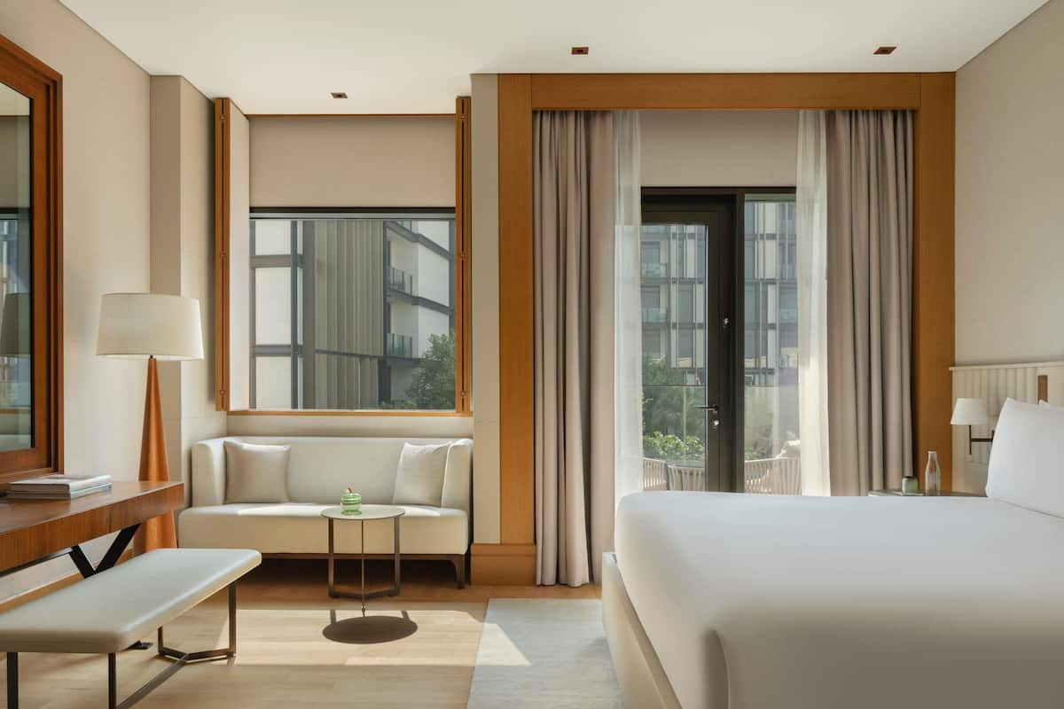 A hotel room with a large bed, a sofa, a small table, a bench, and a window with curtains.