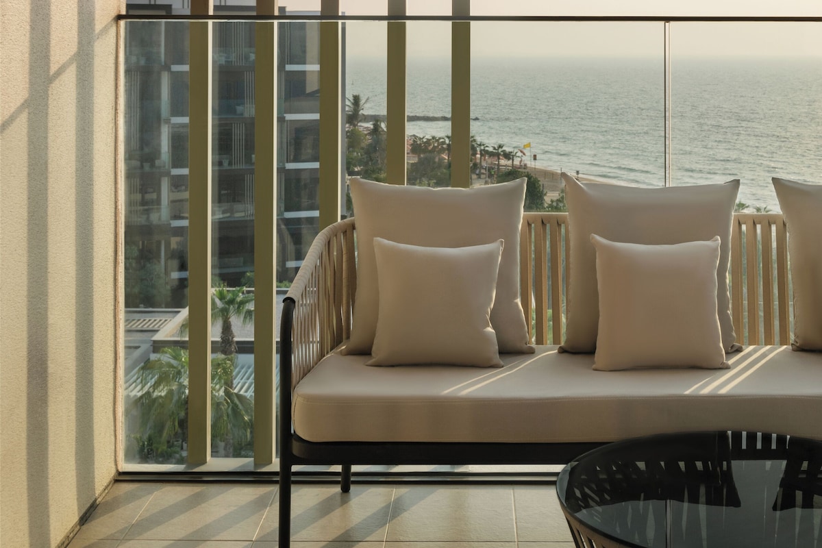 A balcony with a sofa and a glass table, overlooking a body of water and buildings.