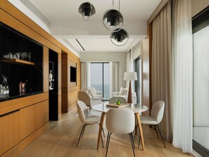 A modern hotel room with a wooden cabinet, a round dining table with chairs, a large window with curtains, and two pendant lights.