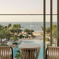 Relax on the balcony with a view of the pool and beach, surrounded by lounge chairs and palm trees.