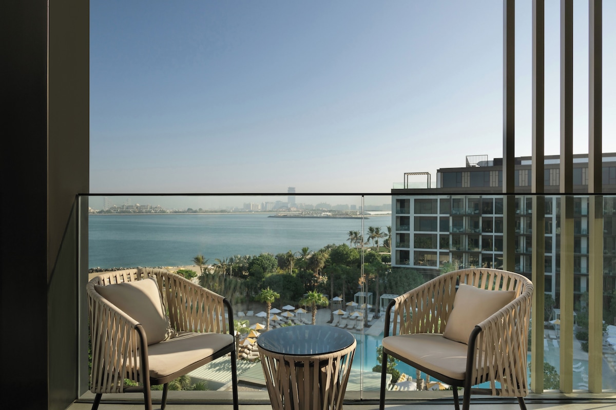 A balcony with wicker chairs and a small table, overlooking a pool and a cityscape with a body of water.