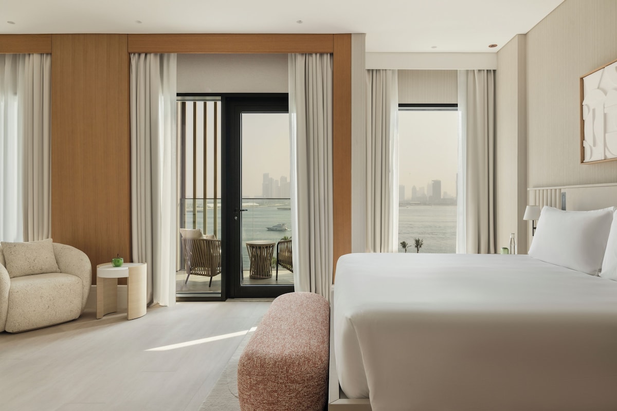 A modern hotel room with a large bed, a seating area with a table, and a view of the city through the window.