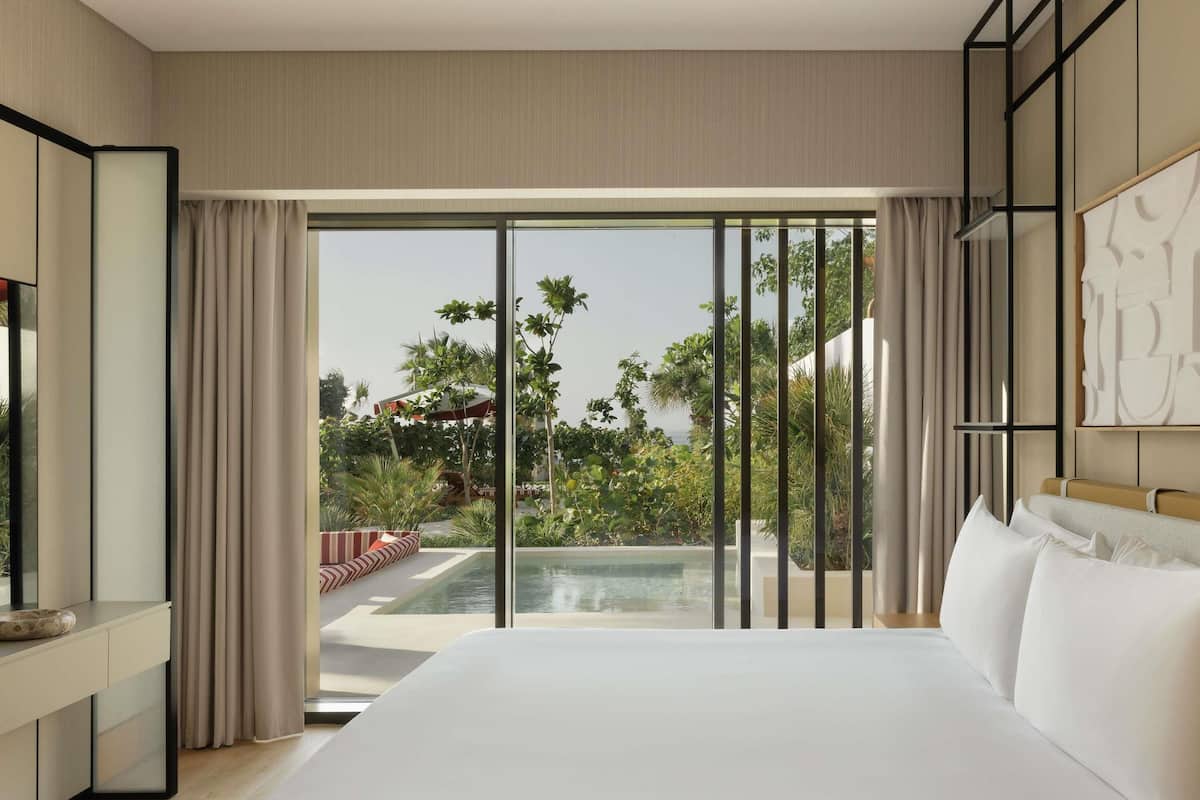 A modern hotel room with a large bed, a TV, a desk, and a view of a pool and greenery.