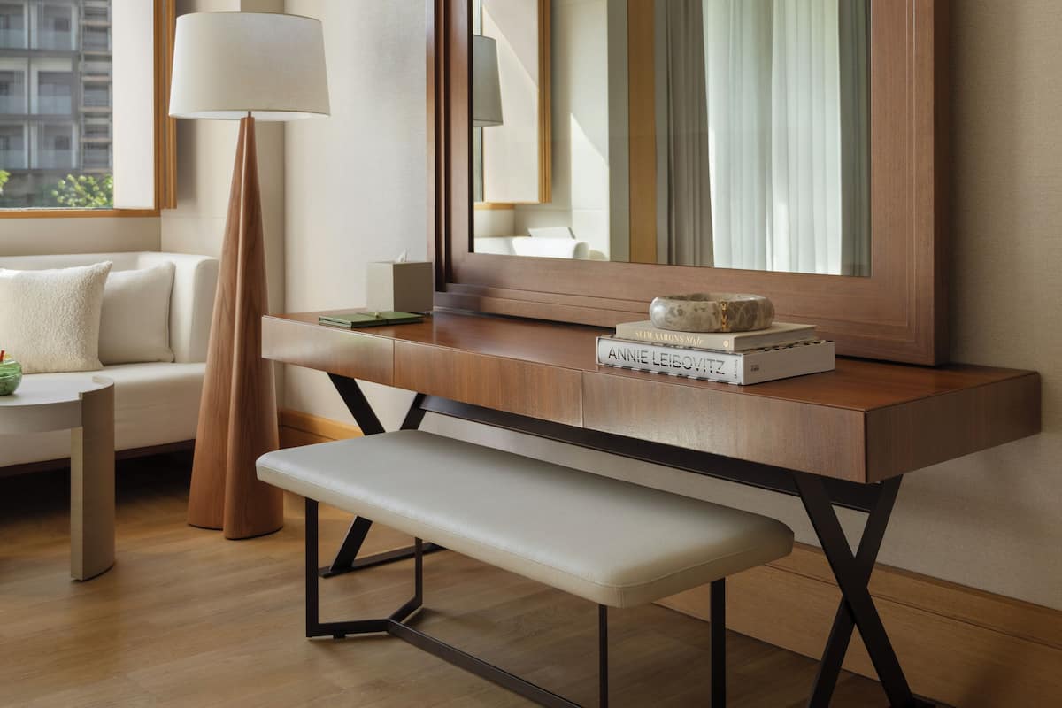 A modern hotel room with a wooden console table, a large mirror, a bench, a floor lamp, and a sofa.