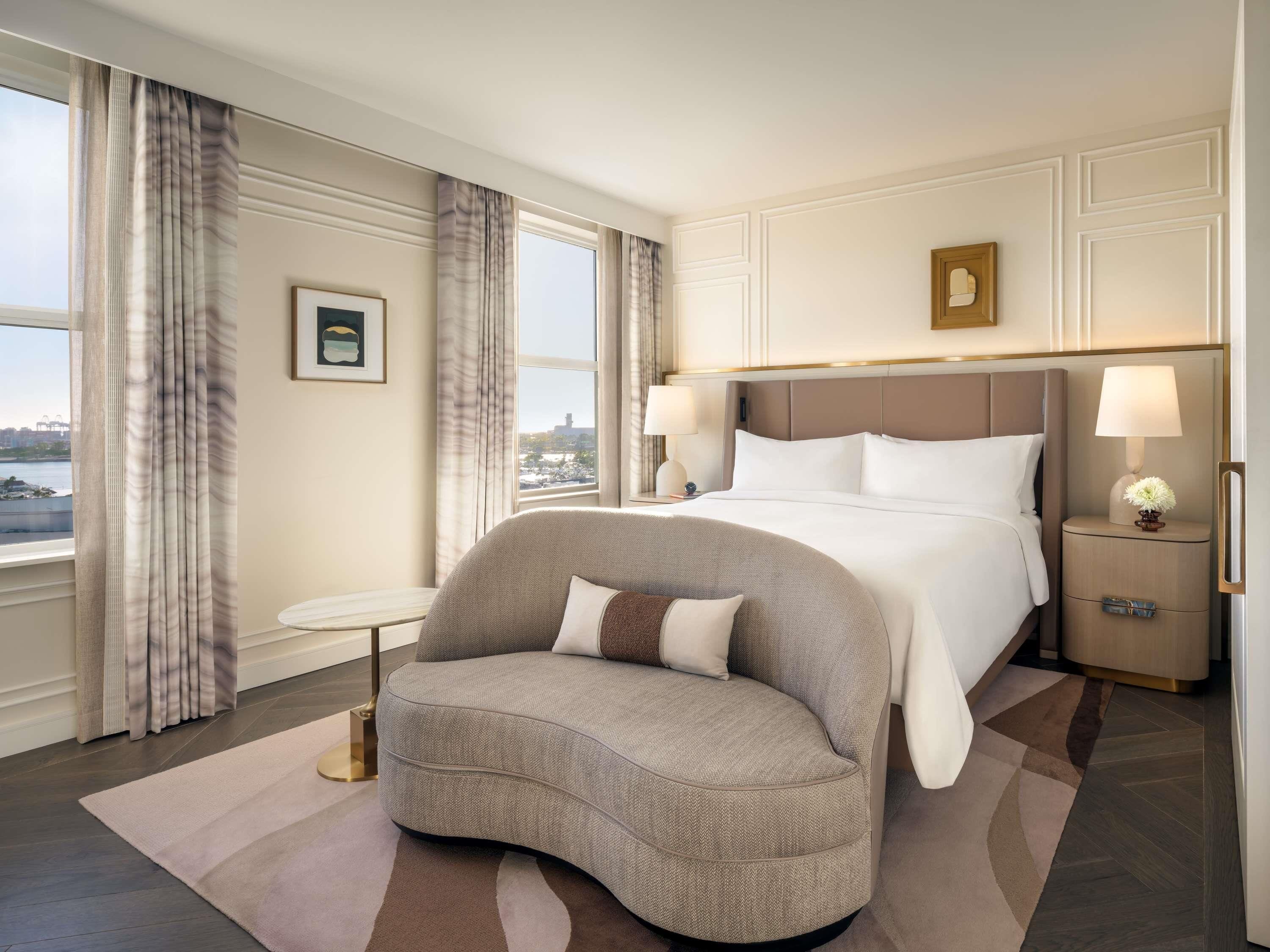 A hotel room with a bed, bedside lamps, a chair, a small table, and a view of the city.