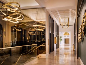 Um saguão de hotel moderno com escadaria de vidro, várias luminárias de cristal e piso de mármore polido.