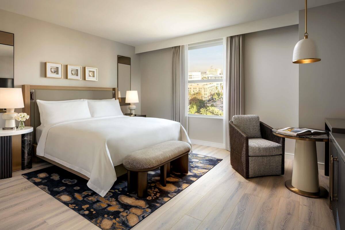 Een hotelkamer met een bed, nachtkastjes, een stoel, een bureau, een lamp, een raam met uitzicht en een hangende lamp.