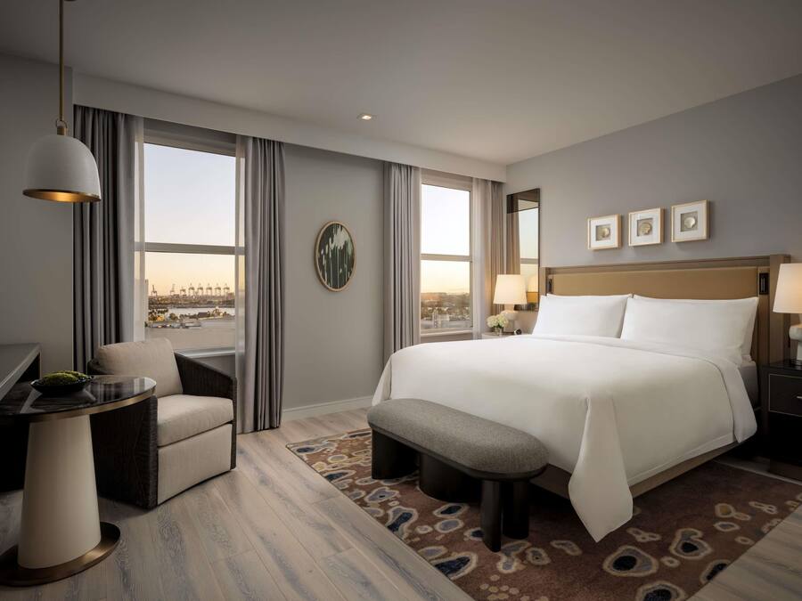 Habitación de hotel con una cama grande, un escritorio, una silla, un banco, vistas de la ciudad y un ventanal amplio.