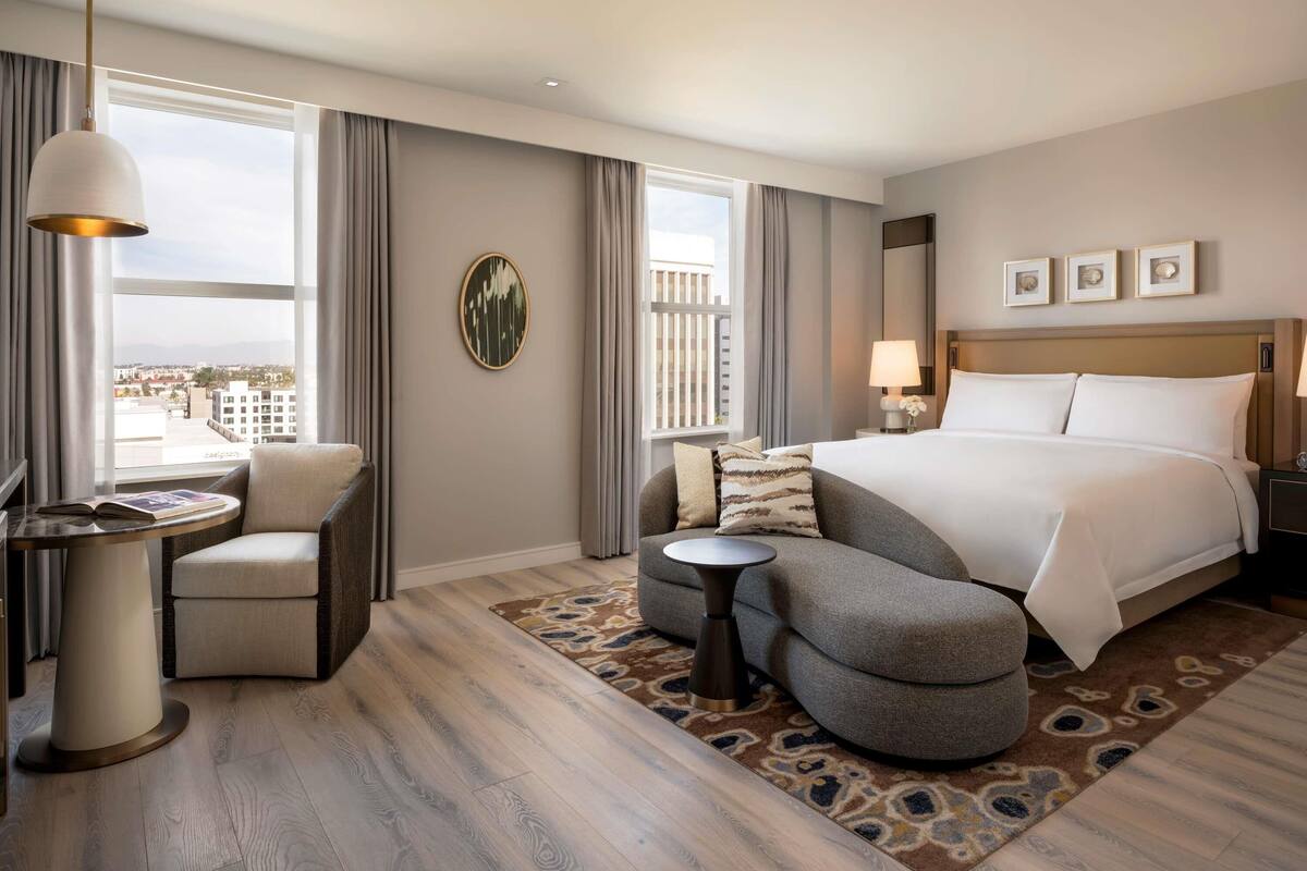 Een moderne hotelkamer met een groot bed, een zithoek met een fauteuil en poef, een tafeltje met een lamp, en uitzicht op de stad door grote ramen.