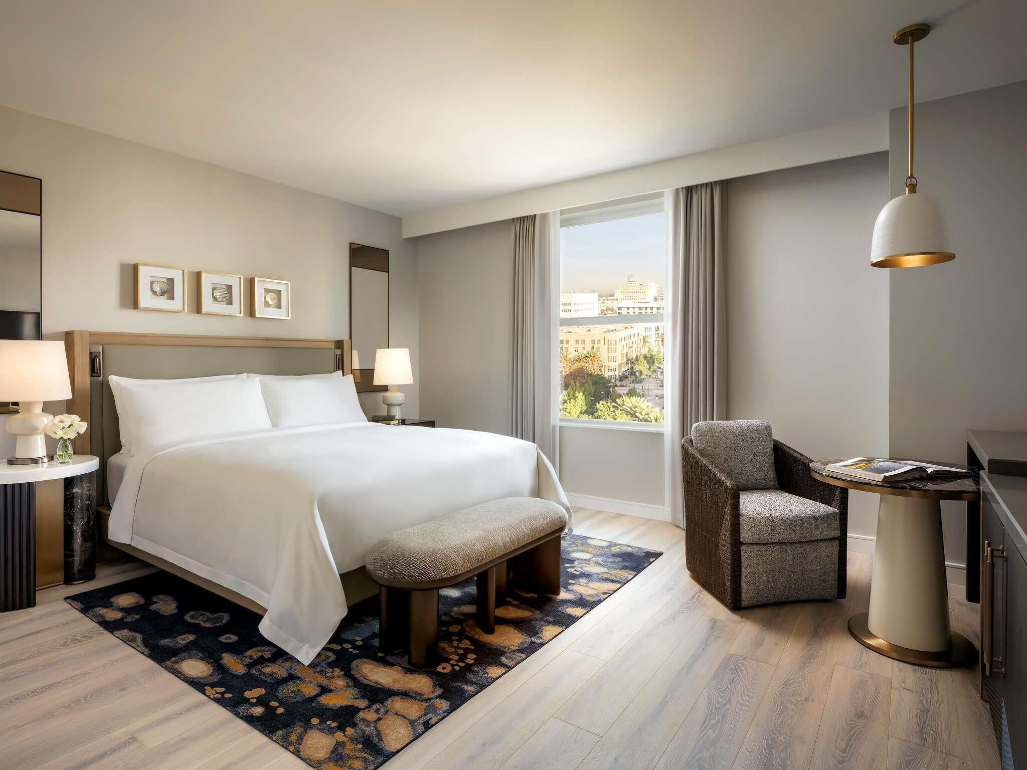 Habitación de hotel con una cama grande, una silla, un escritorio y vistas a la ciudad a través de la ventana.