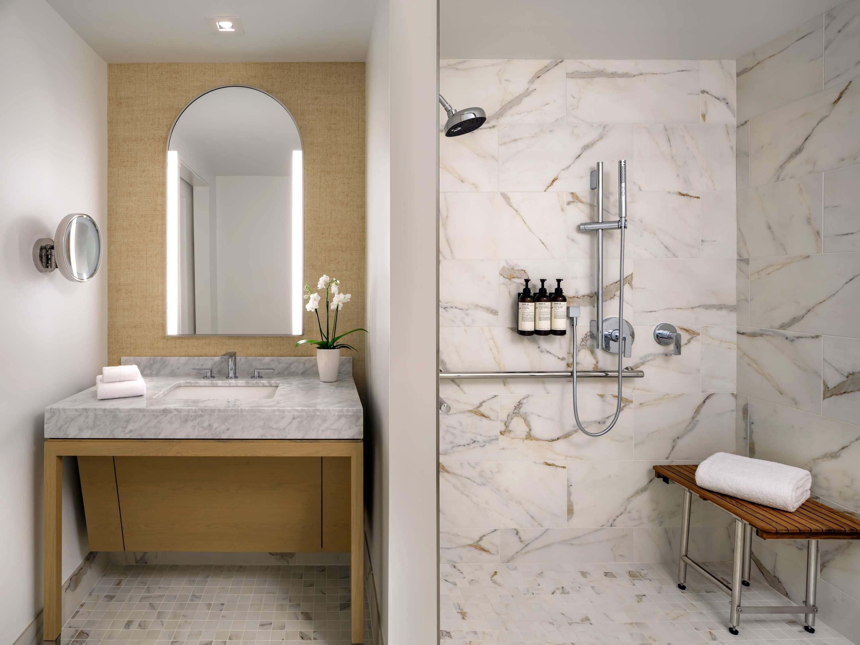 A bathroom with a marble sink, a mirror, a potted orchid, a shower with a handheld showerhead, and a bench with a towel.