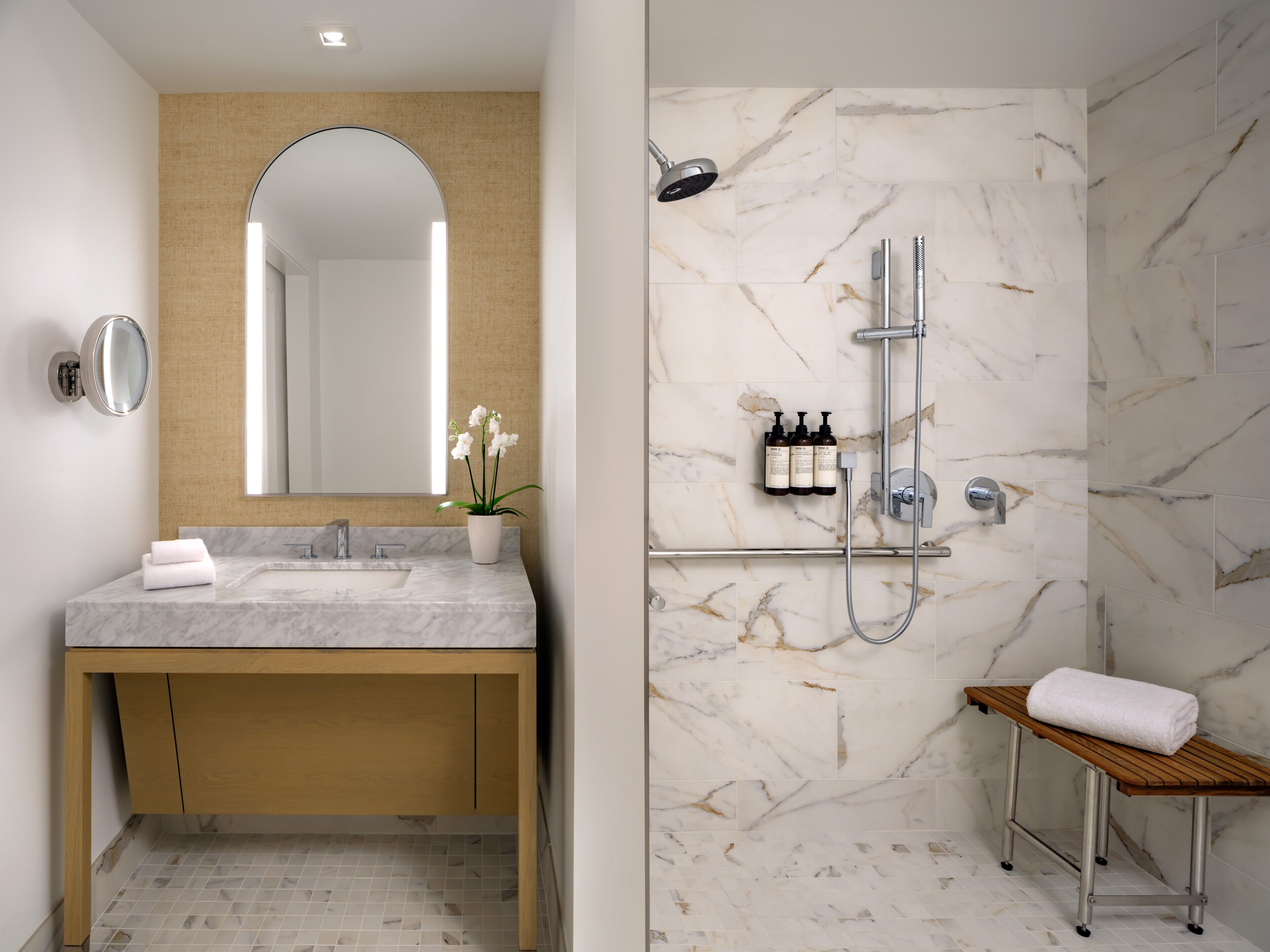Un baño moderno con tocador de mármol, un espejo grande, ducha con cabezal de ducha manual y un banco.