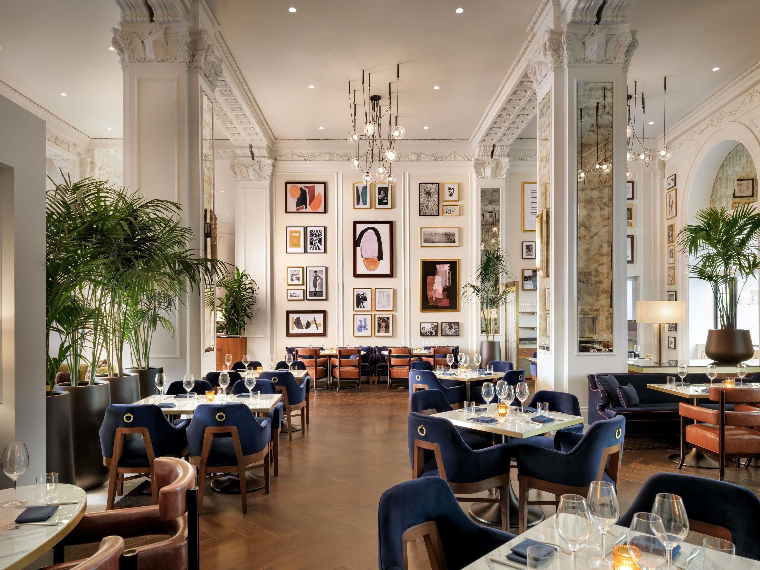 A spacious dining area with high ceilings, ornate columns, and a variety of framed artwork on the walls. Tables are set with chairs, and there are potted plants adding greenery.