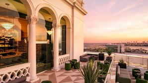Un lounge en la azotea con vistas a la ciudad al atardecer, que incluye una zona de bar, asientos y columnas decorativas.