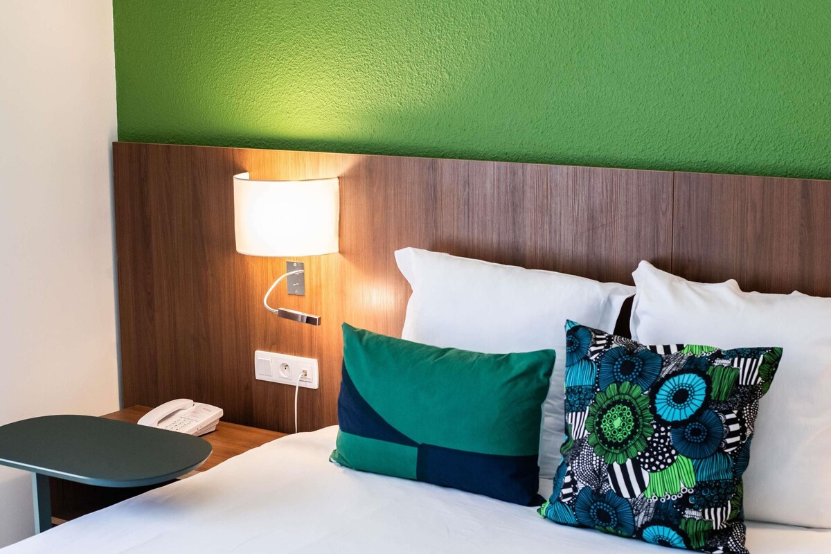 A hotel room with a wooden headboard, a bedside lamp, a telephone, and a bed with green and patterned pillows.