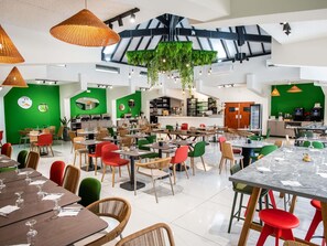 A spacious dining area with multiple tables and chairs, a kitchen area with bar seating, and hanging plants. - Ibis Styles Cayenne Centre Amazonia (Ouverture Juin 2024) (Cayenne)