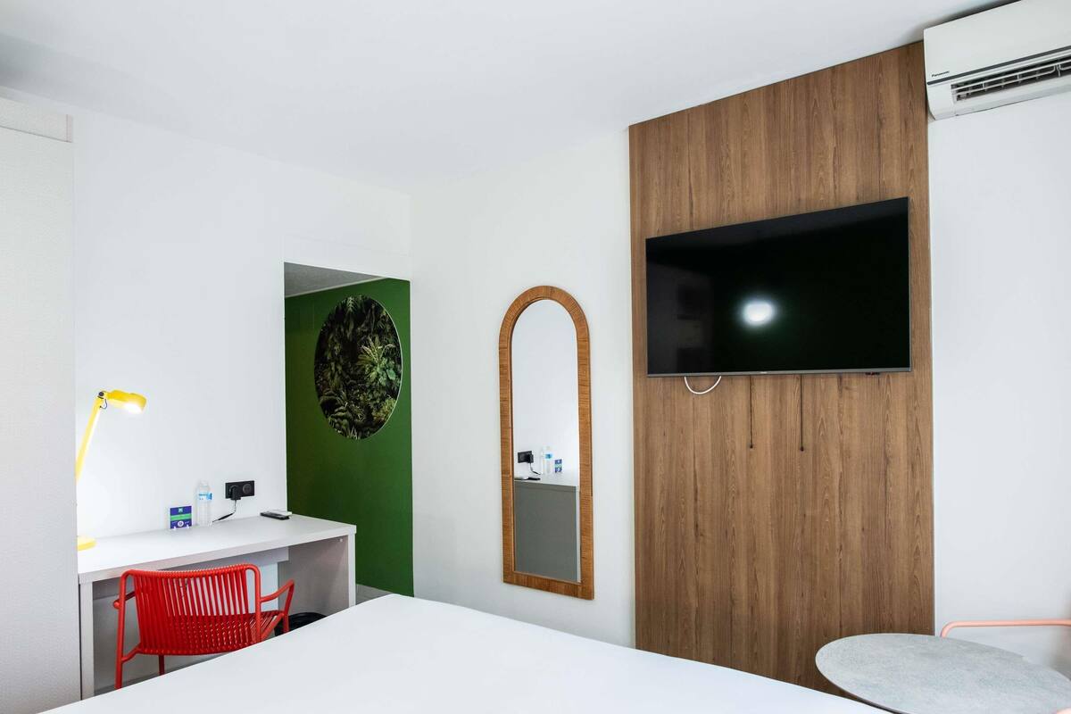 A hotel room with a bed, a desk with a chair, a TV mounted on the wall, and an air conditioning unit.