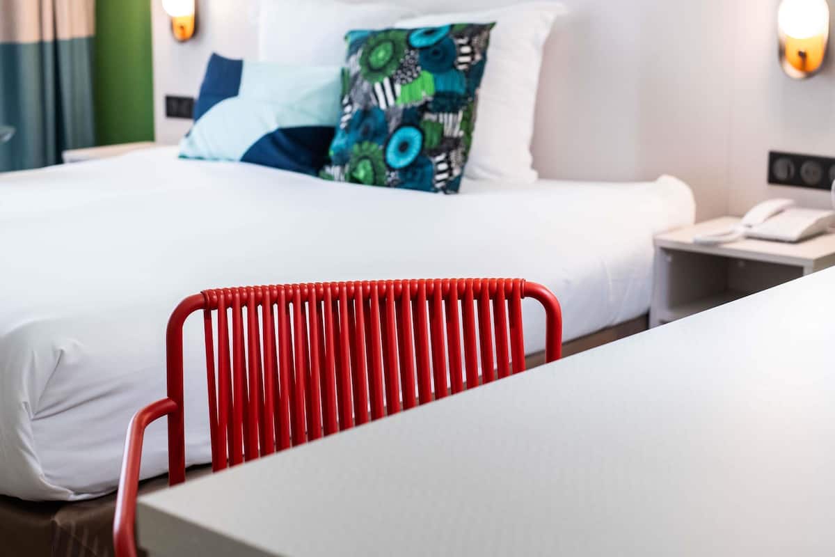 A hotel room with a bed, a red chair, a bedside table with a phone, and a wall-mounted light.