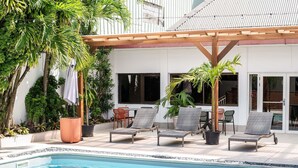 Zona junto a la piscina con sillones de descanso, una pérgola y plantas en macetas.