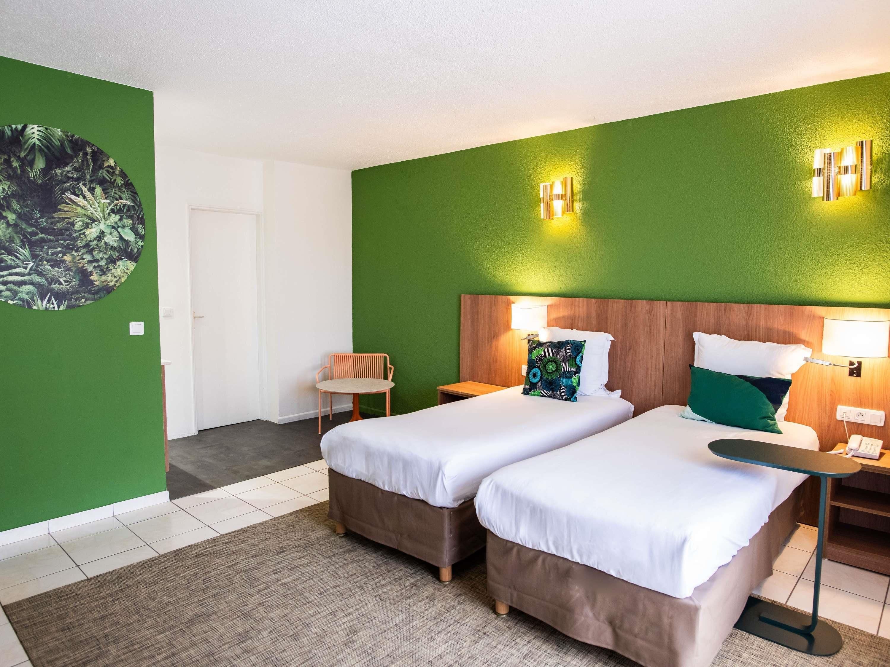 Une chambre d’hôtel avec deux lits, un mur d’accent vert, un miroir rond et une petite table.