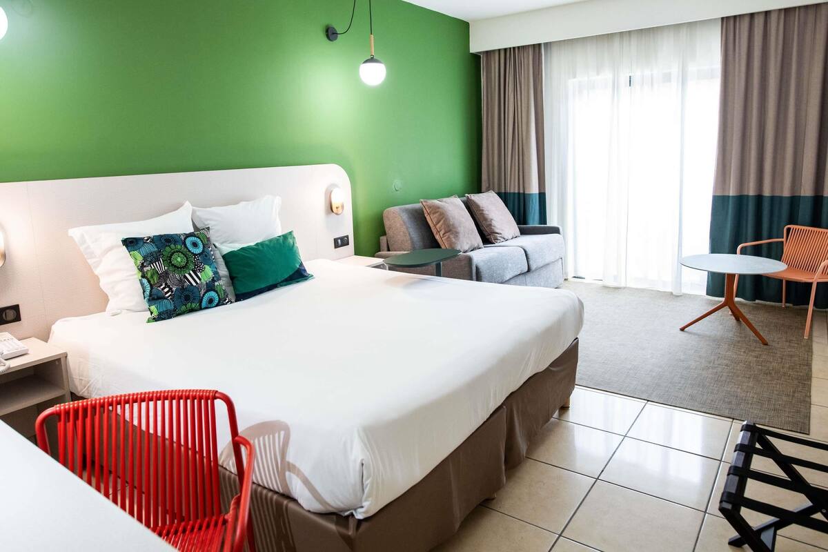 A hotel room with a bed, a desk, a red chair, a grey sofa, and a round table.